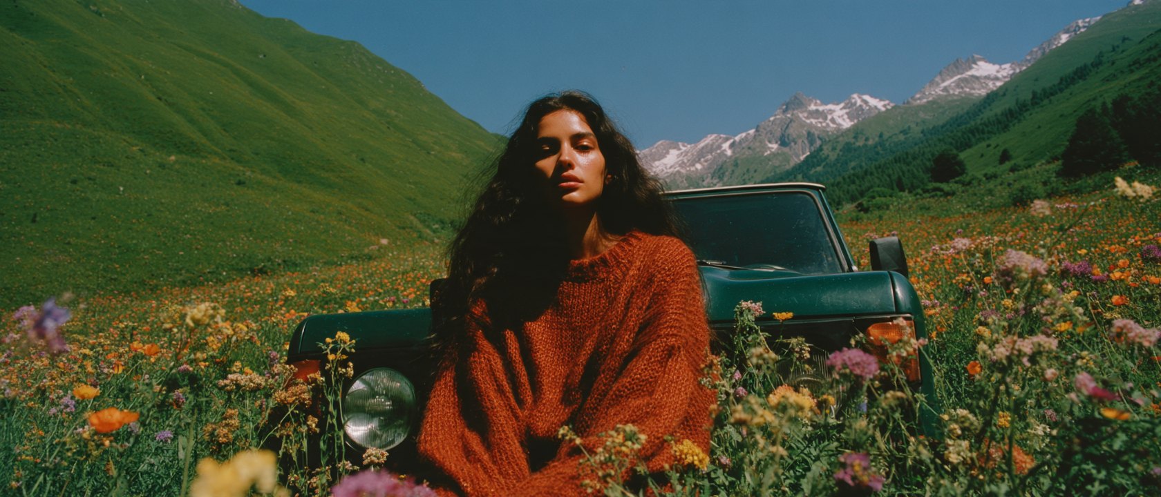Woman in a rust sweater sitting against a dark green SUV in an alpine wildflower meadow