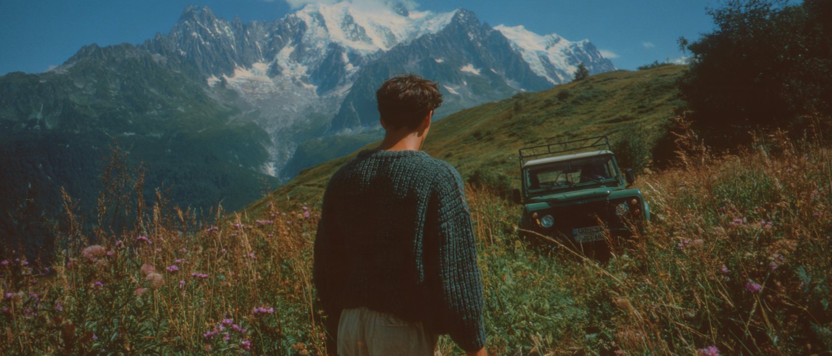 Man walking through a meadow toward his car below snow peaks